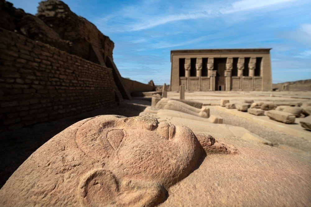 A weathered carved face in the foreground with a Dendera temple building behind