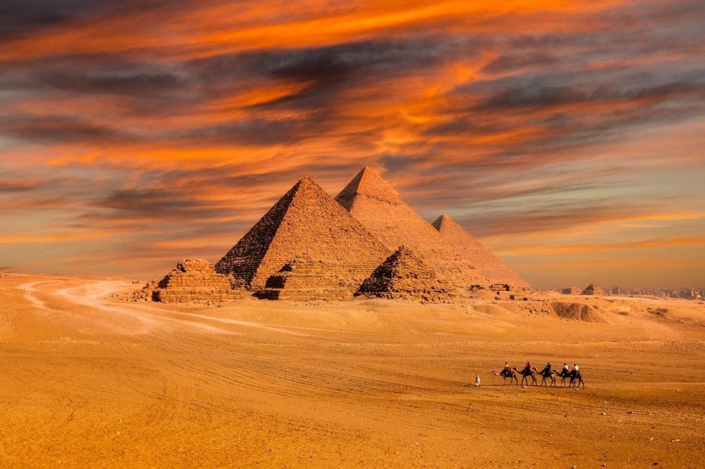 The Pyramids of Giza under an orange sunset sky with camels crossing the sand