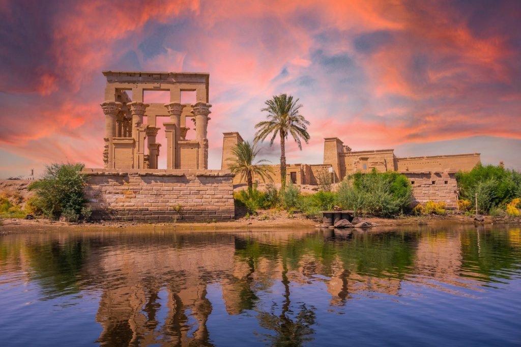 The Temple of Philae and its kiosk reflected in the water under an orange sunrise sky