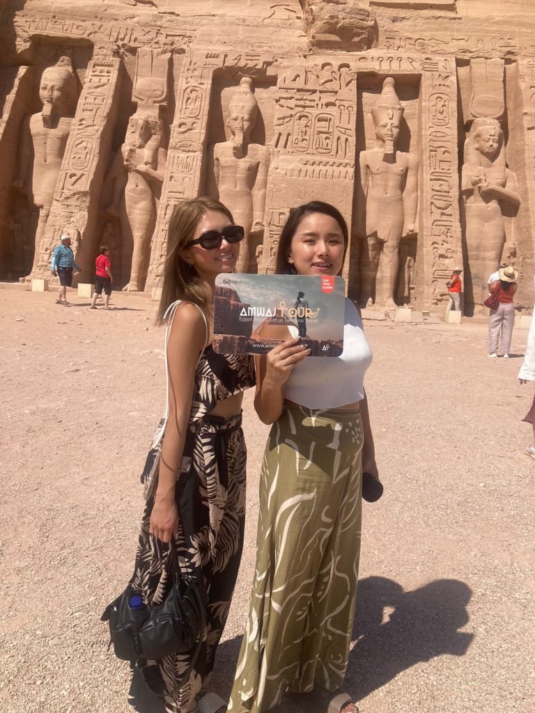 Two travelers with an Amwaj Tour photo card in front of the Temple of Nefertari at Abu Simbel