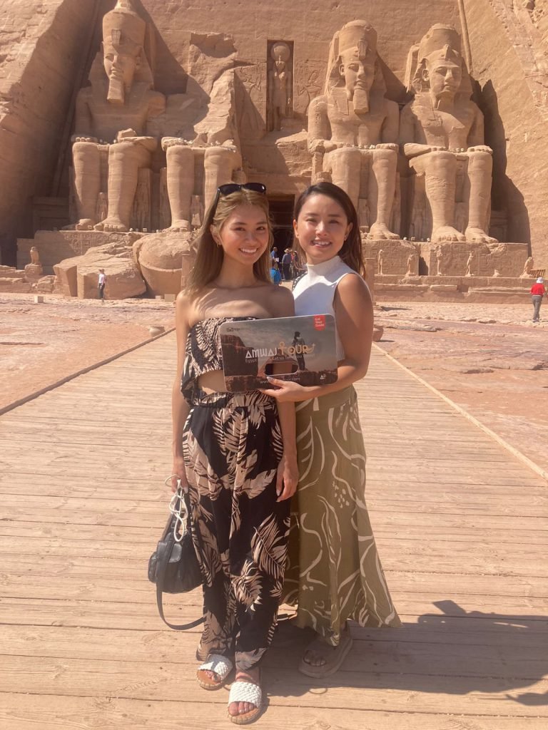 Two travelers with an Amwaj Tour photo card in front of the four colossi of the Great Temple of Abu Simbel