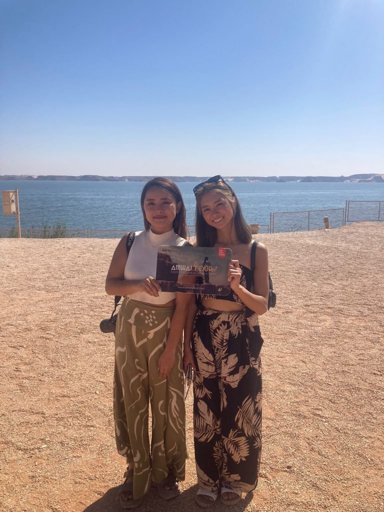 Two travelers with an Amwaj Tour photo card on the shore of Lake Nasser at Abu Simbel