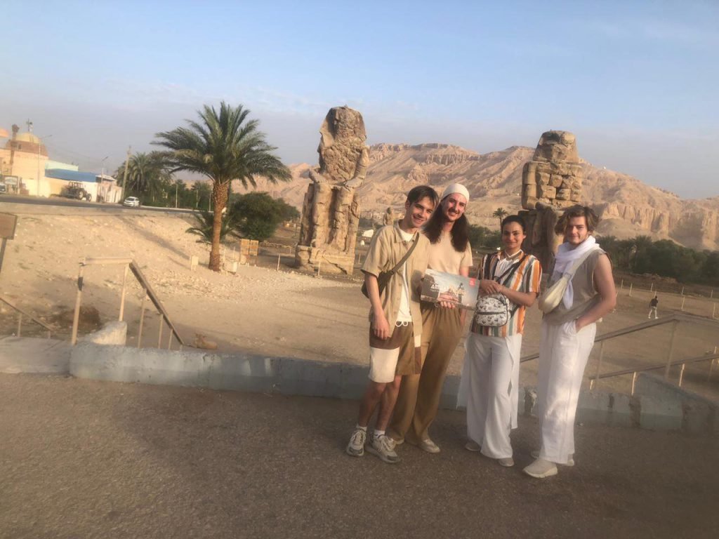 Four friends with an Amwaj Tour sign in front of the Colossi of Memnon in Luxor at golden hour