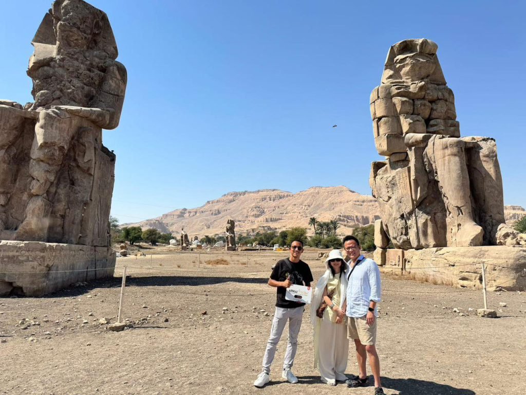 Three travelers with an Amwaj Tour sign between the Colossi of Memnon