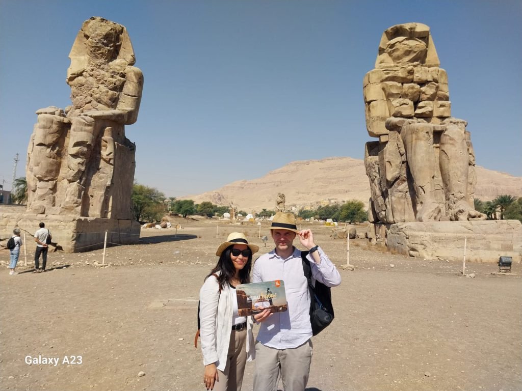A couple with an Amwaj Tour photo card in front of the Colossi of Memnon