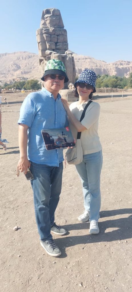 A couple with an Amwaj Tour photo card in front of a Colossus of Memnon