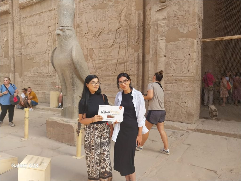 Two travelers holding an Amwaj Tour sign beside the granite falcon of Horus at Edfu Temple
