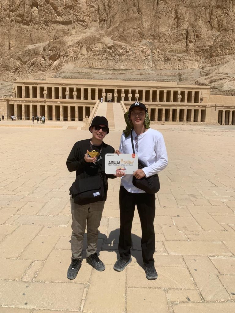 Two travelers with an Amwaj Tour sign in front of the Temple of Hatshepsut