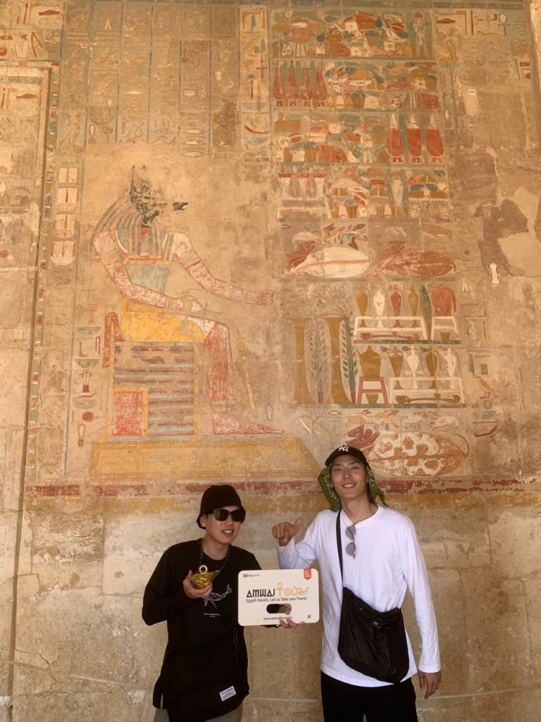 Two travelers with an Amwaj Tour sign under a painted relief in the Temple of Hatshepsut