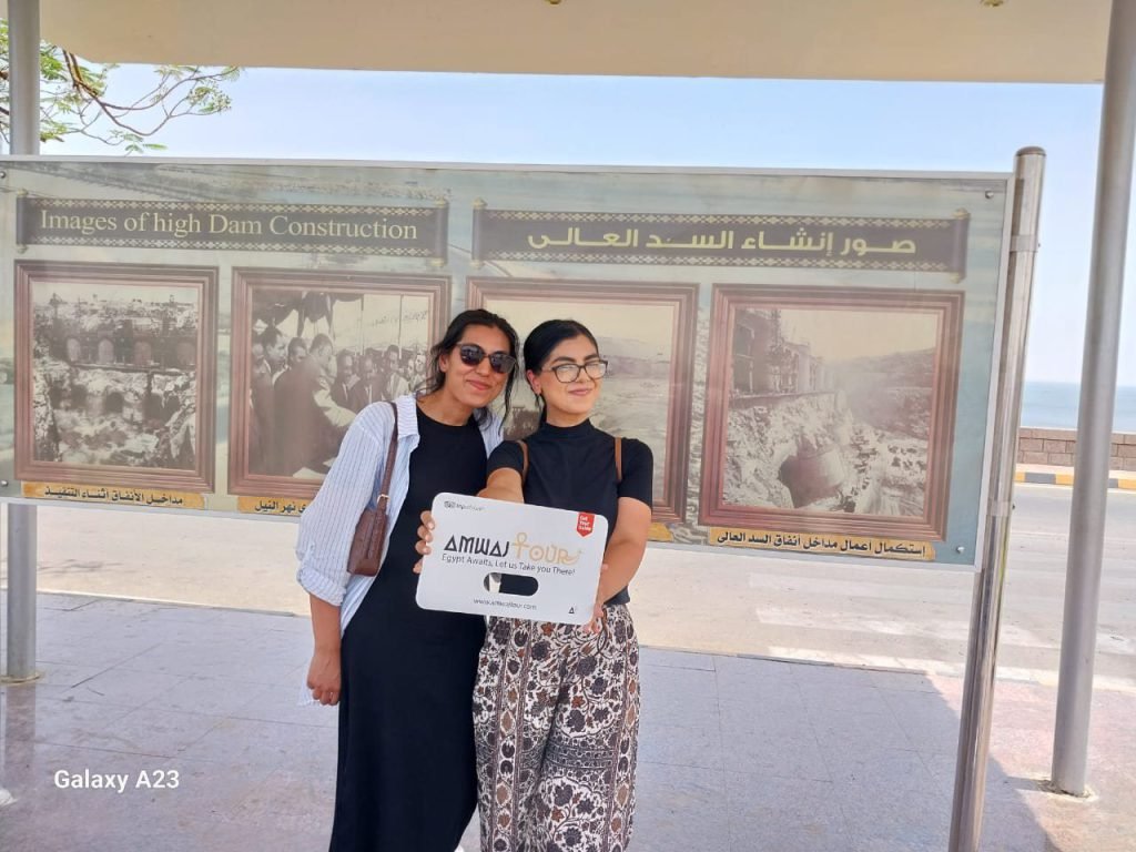 Two travelers with an Amwaj Tour sign by the historic photo boards at the Aswan High Dam