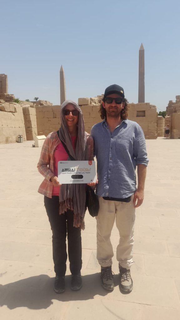 A couple with an Amwaj Tour sign in front of the obelisks of Karnak Temple
