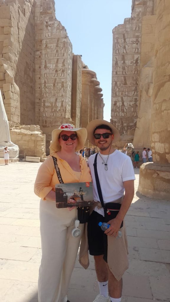 A couple in sun hats with an Amwaj Tour photo card in the great court of Karnak Temple