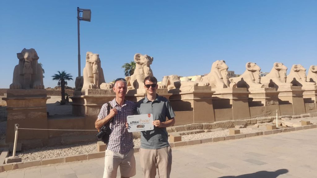 Two travelers with an Amwaj Tour sign on the avenue of ram-headed sphinxes at Karnak Temple