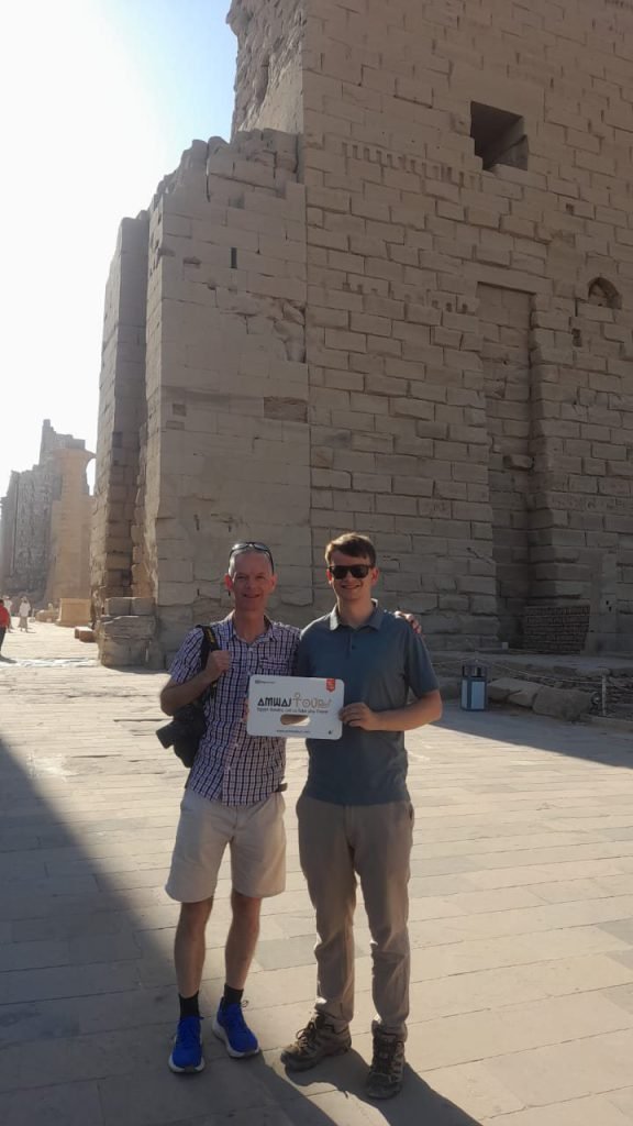 Two travelers with an Amwaj Tour sign in front of the great gateway of Karnak Temple