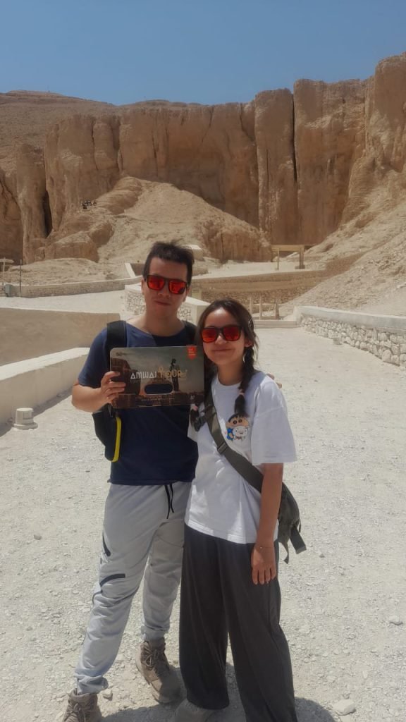 A couple with an Amwaj Tour photo card below the cliffs of the Theban hills