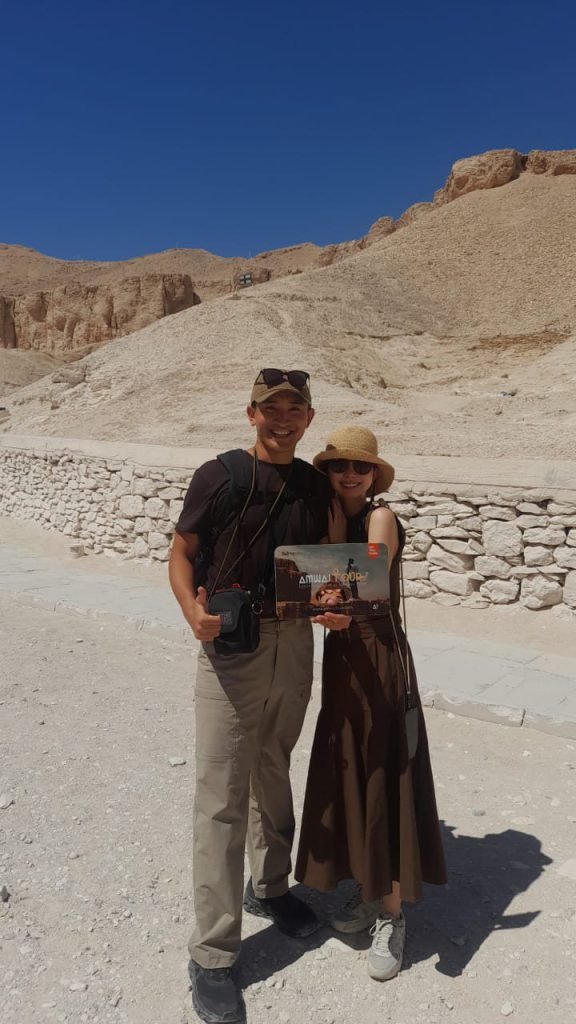 A couple with an Amwaj Tour photo card in the Valley of the Kings under a deep blue sky