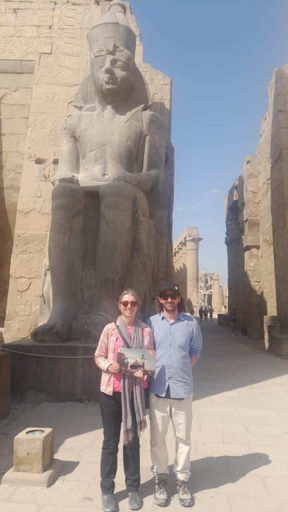 A couple with an Amwaj Tour photo card beside the seated statue of Ramses II at Luxor Temple