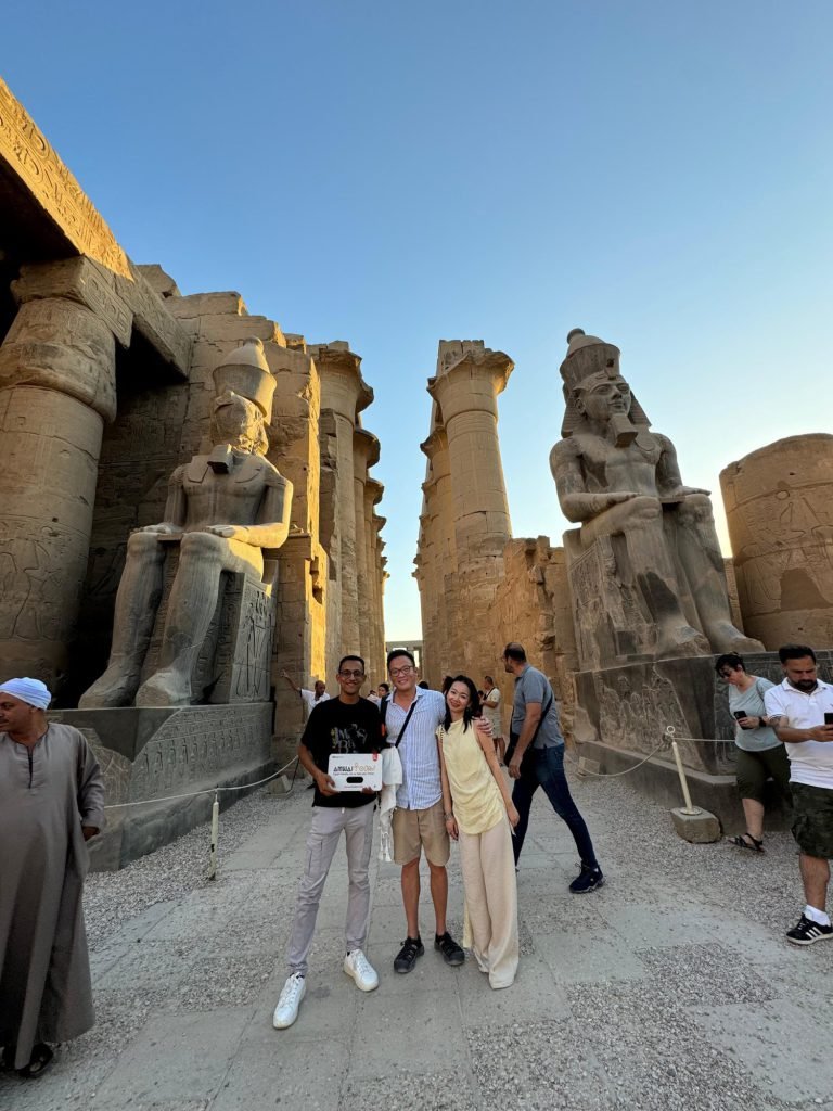 A group of travelers with an Amwaj Tour sign before the seated statues of Ramses II at Luxor Temple