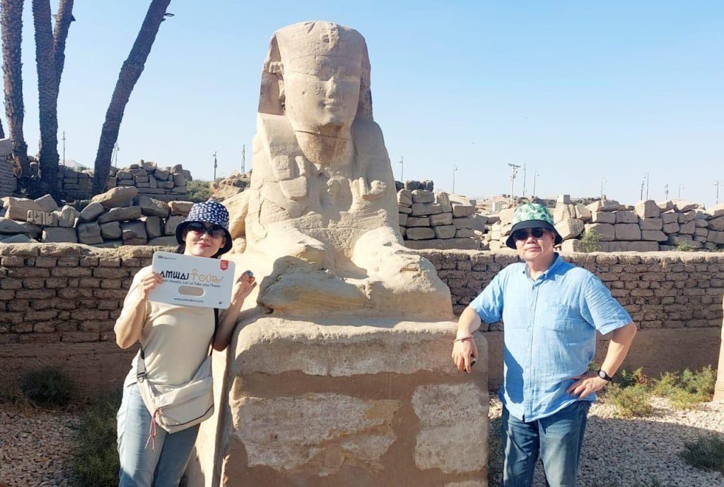 Two travelers with an Amwaj Tour sign beside a sphinx on the Avenue of Sphinxes in Luxor