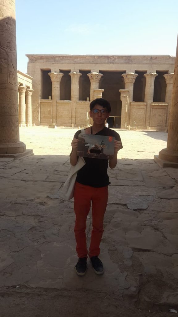 A traveler with an Amwaj Tour photo card in a colonnaded temple courtyard at Philae