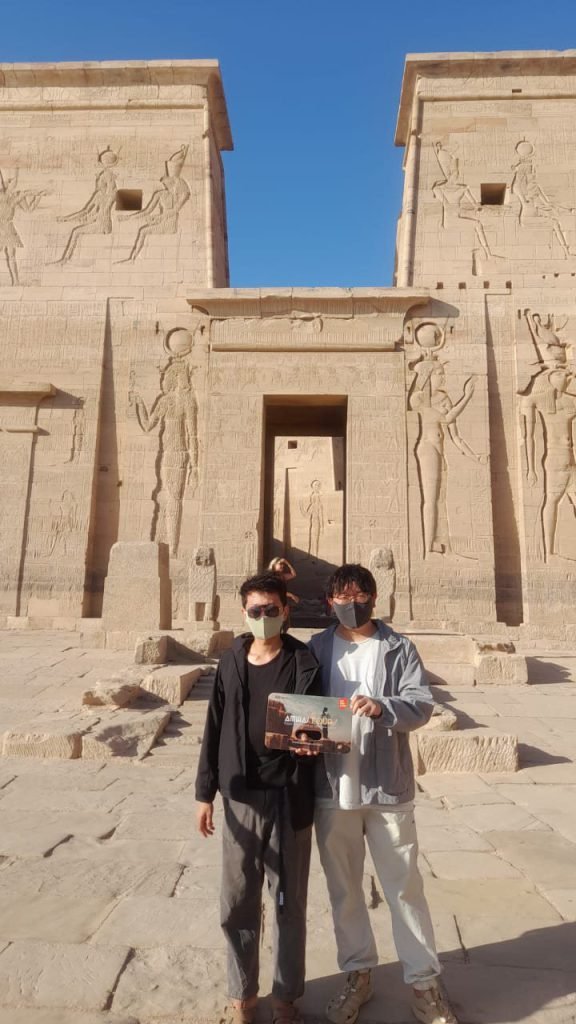 Two travelers with an Amwaj Tour photo card at a carved gateway of Philae Temple