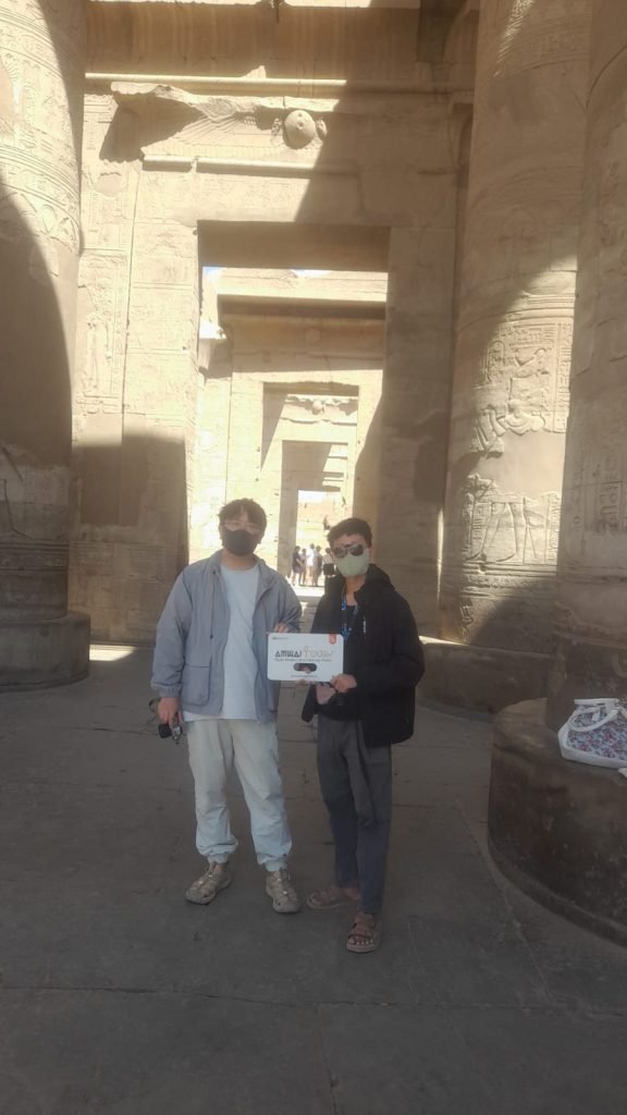 Two travelers with an Amwaj Tour sign in a shaded stone passage of an ancient temple